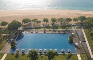 Vista aérea de un resort con una amplia piscina rectangular rodeada de sillas de descanso y sombrillas, una playa de arena al fondo y exuberante vegetación.