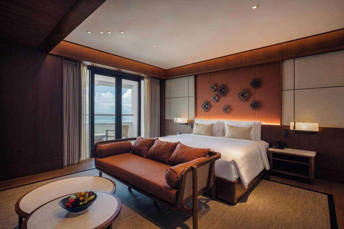 A hotel room with a large bed, a sofa, a small table with fruit, and a view of the ocean.