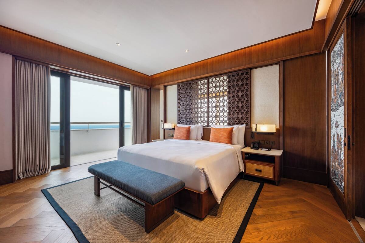 A hotel room with a large bed, a bench, a nightstand, a lamp, and a view of the sea through a sliding door.