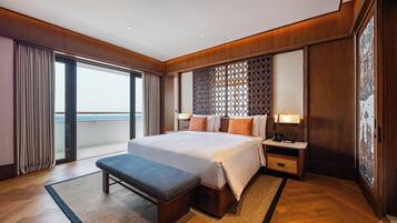 Een hotelkamer met een groot bed, een bankje, een nachtkastje, een lamp en uitzicht op de zee door een schuifdeur.