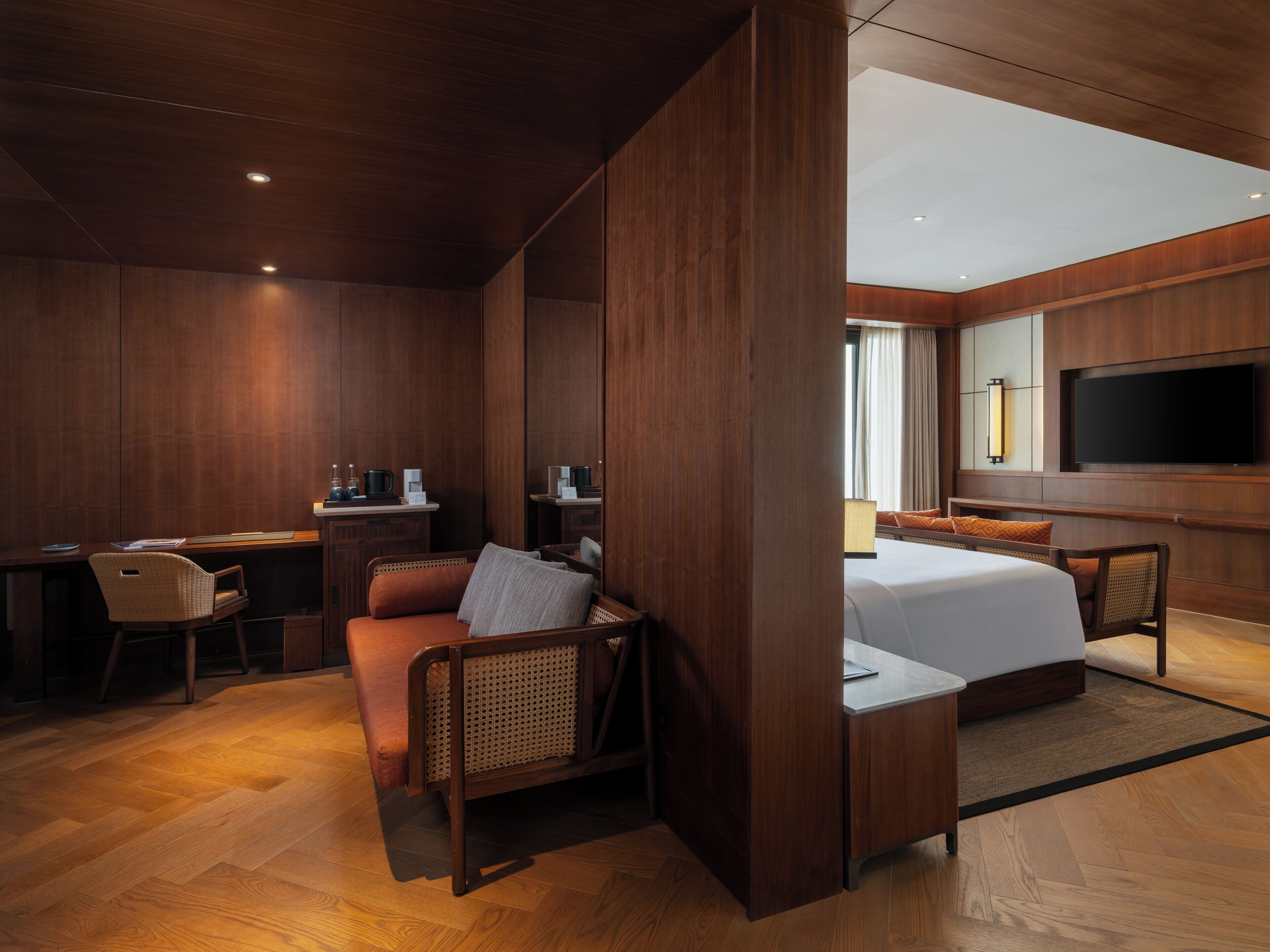 A hotel room with a wooden interior, a bed, a desk with a chair, a sofa, and a TV mounted on the wall.