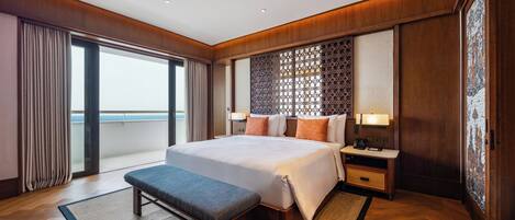 A spacious hotel room with a large bed, a bench, a nightstand, and a view of the sea through a sliding door.