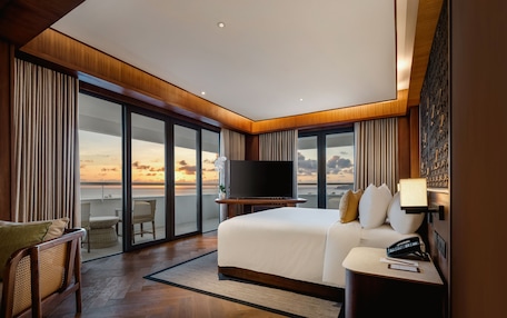 A modern hotel room with a large bed, a flat-screen TV, a bedside table, and a view of the sunset through floor-to-ceiling windows.