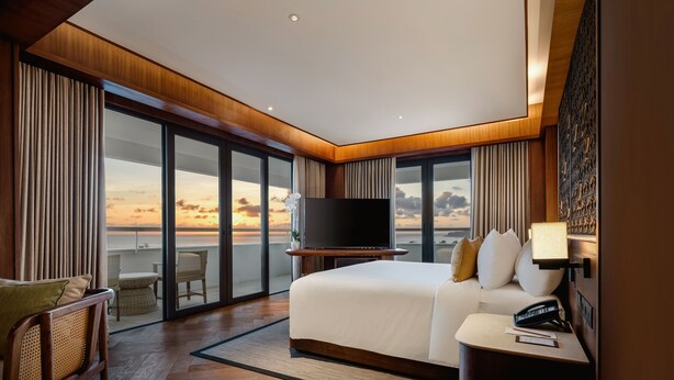 Una habitación de hotel moderna con una cama grande, un televisor de pantalla plana, una mesita de noche y vistas al atardecer a través de ventanales de gran altura.