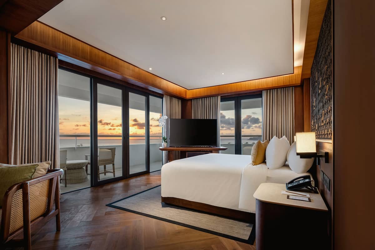 A modern hotel room with a large bed, a flat-screen TV, a bedside table, and a view of the sunset through floor-to-ceiling windows.