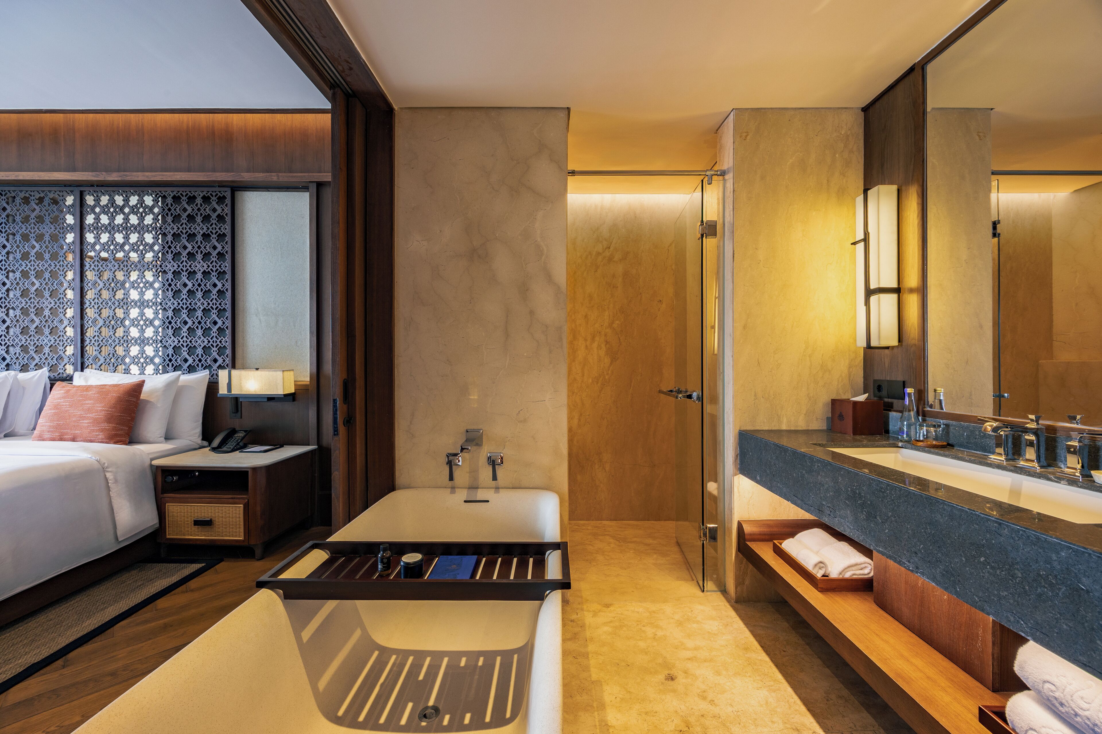 A hotel room with a bed, bedside table, lamp, and a large mirror above the sink.