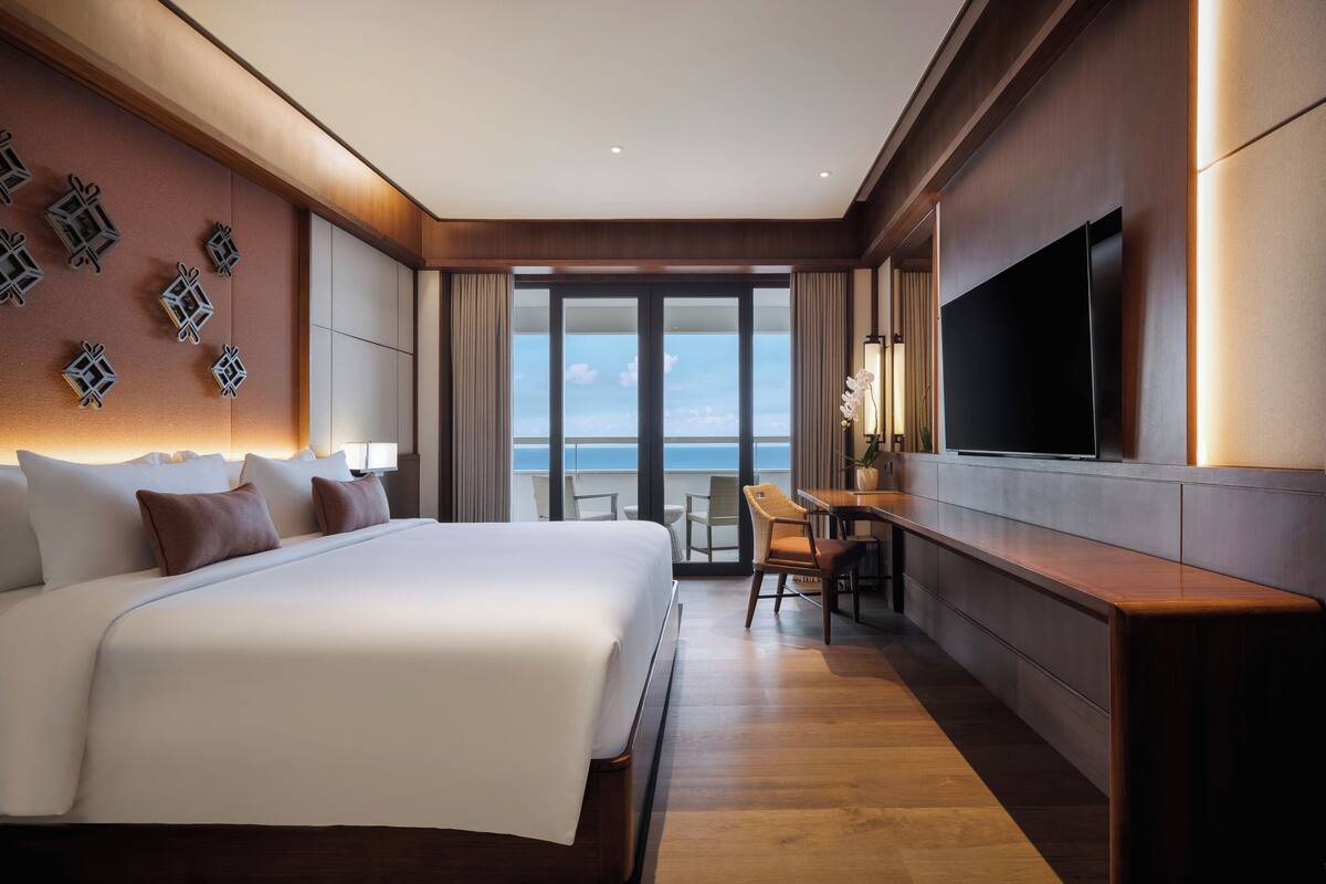 A hotel room with a large bed, a desk, a flat-screen TV, and a view of the ocean.
