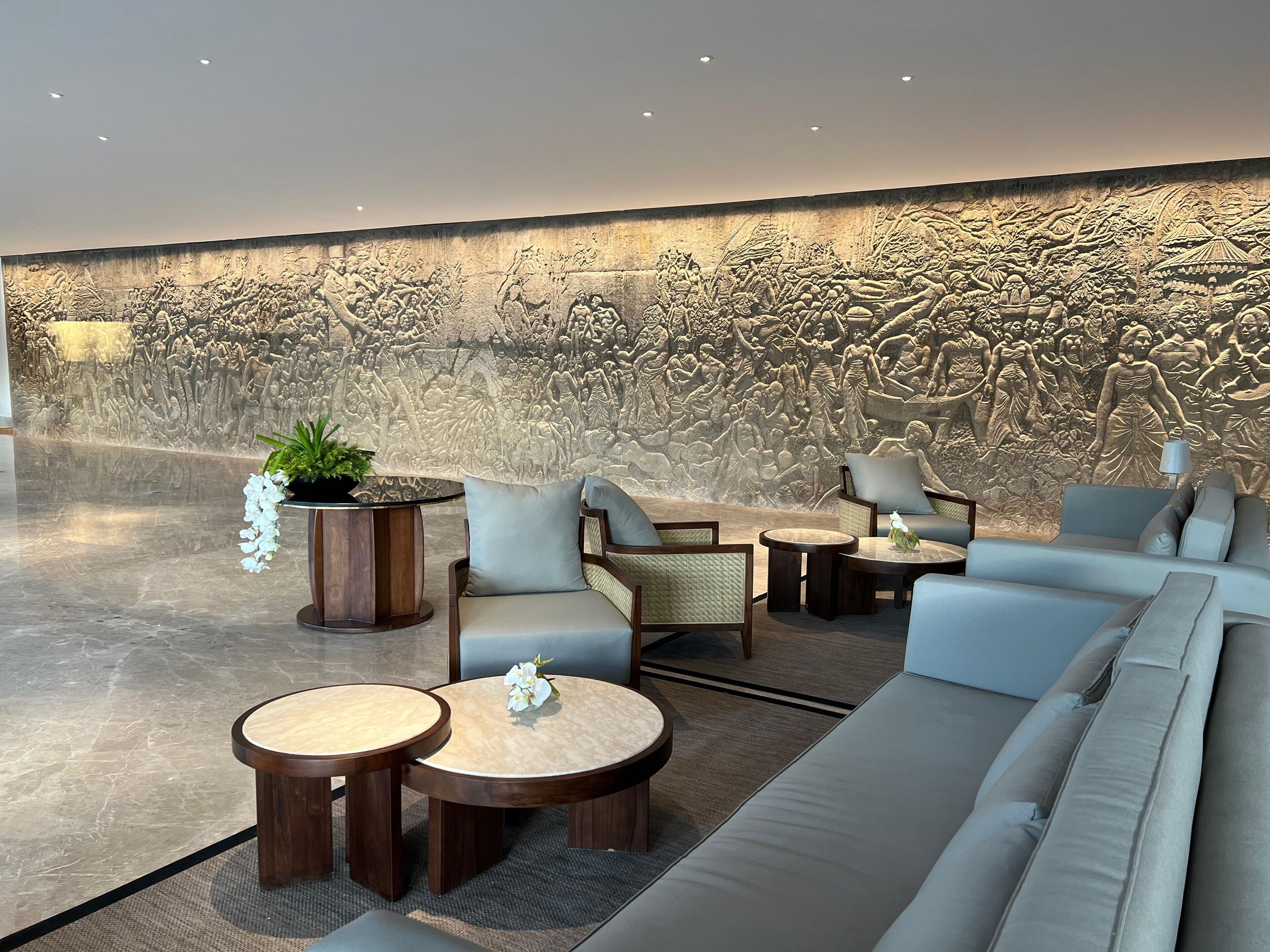 Eine Lobby mit einer großen, detaillierten Reliefwand, einer Sitzecke mit Sofas und runden Tischen sowie einer Topfpflanze.
