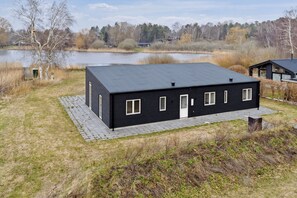 A black building with a flat roof, a paved walkway, and a body of water in the background. - Brand new and modern summer house in Kulhuse (Jægerspris)