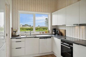 A modern kitchen with white cabinets, a dishwasher, an oven, and a window with a view of greenery.