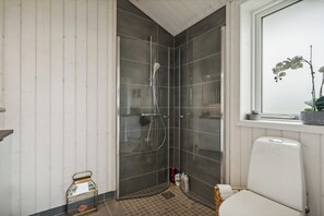 A bathroom with a glass shower enclosure, a white toilet, a window with a potted orchid, and a small trash bin.