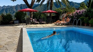 A swimming pool with a person swimming, surrounded by lounge chairs and red umbrellas, with mountains in the background. - Ferienapartment im Sonnigen Westen von La Palma (El Paso)