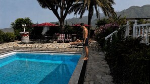 En swimmingpool med flisekant, omgivet af liggestole og parasoller, med et bjergrigt landskab i baggrunden.