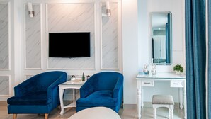 Quarto de hotel moderno com televisão de tela plana na parede, uma escrivaninha branca com espelho, duas poltronas azuis e um banquinho pequeno.