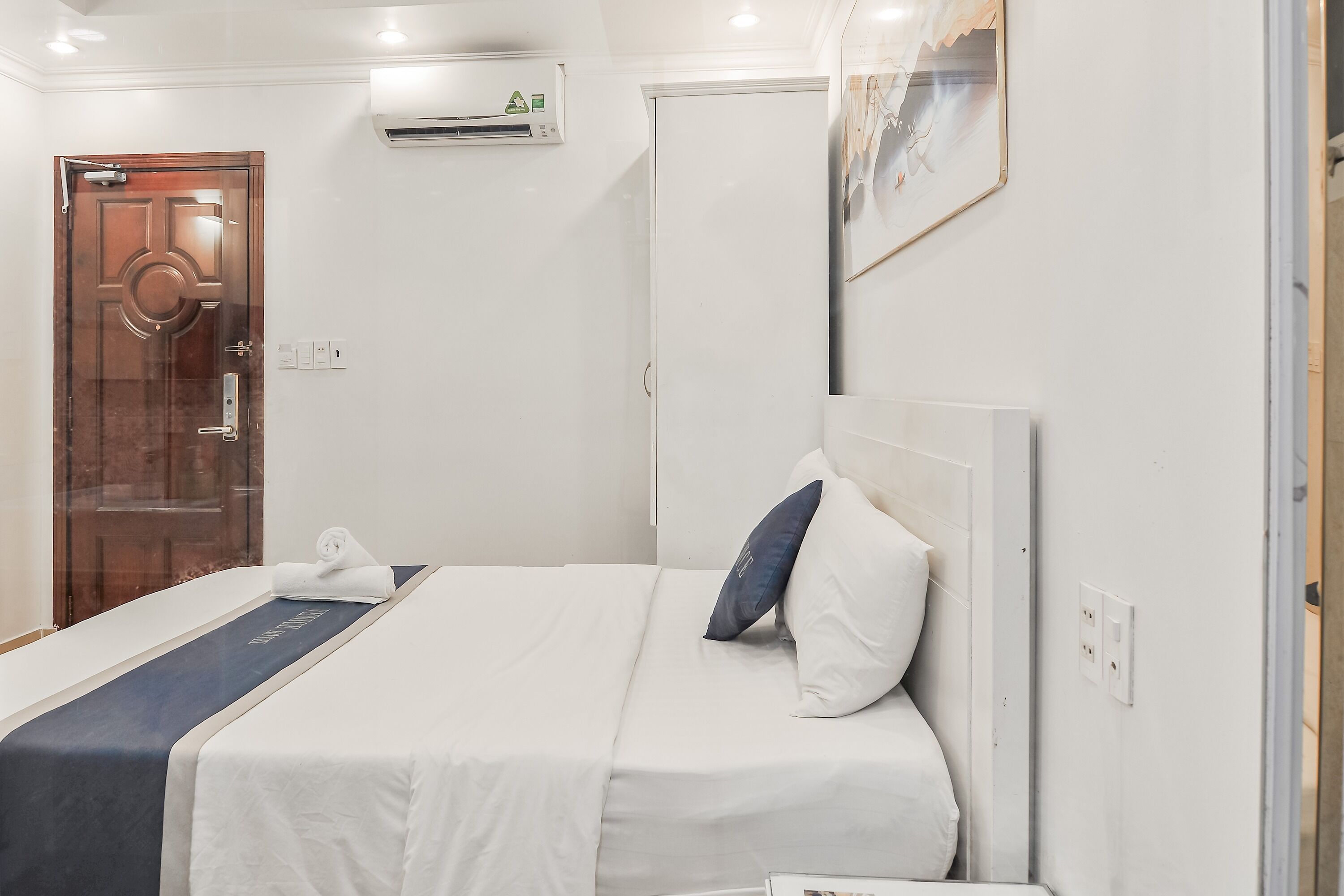 A hotel room with a bed, a wooden door, an air conditioning unit, and a painting on the wall.