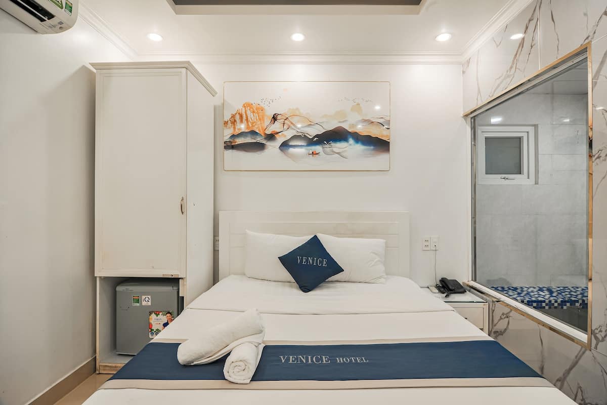 Une chambre d’hôtel comprenant un lit, une table de chevet, une petite armoire, un tableau et une salle de bain avec un miroir et des carreaux.