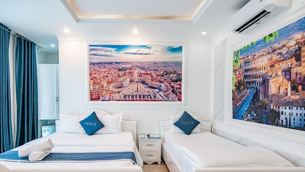 Quarto de hotel com duas camas, uma grande pintura de paisagem urbana acima das camas e vista de Veneza nos travesseiros.