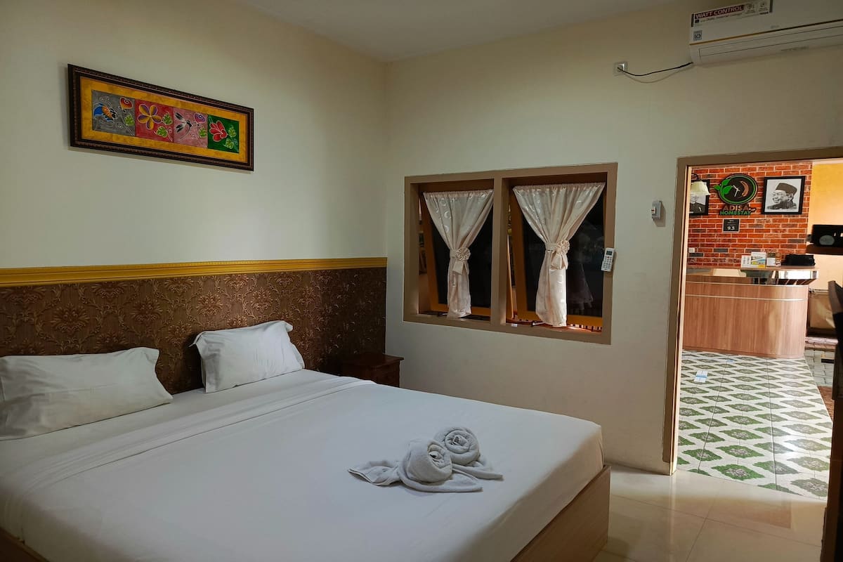 A hotel room with a neatly made bed, a framed artwork on the wall, and a view of the kitchen area through an open door.