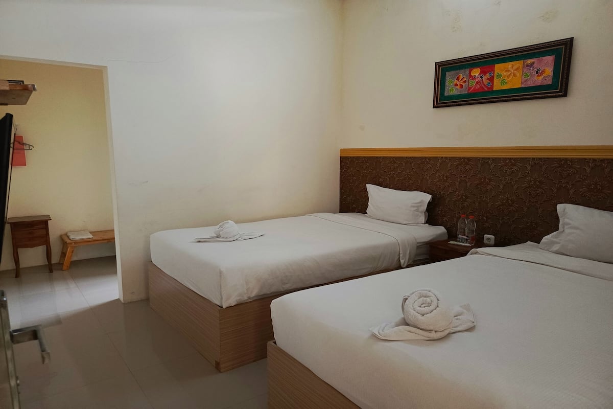 A hotel room with two beds, a wooden headboard, a small bedside table, a wall-mounted painting, and a door leading to another room.