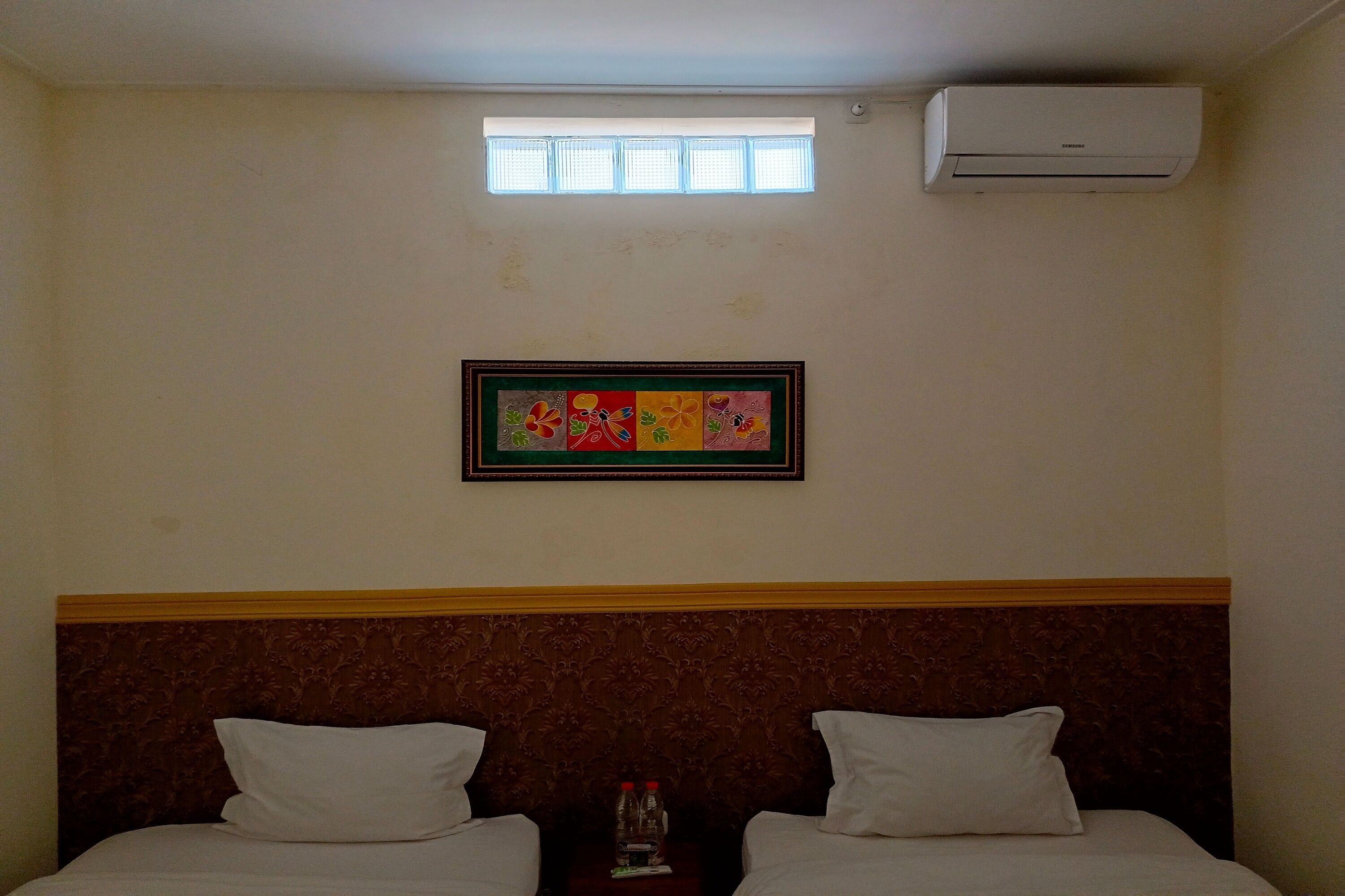 Ein Hotelzimmer mit zwei Betten, einem gerahmten Kunstwerk an der Wand, einer Klimaanlage und einem kleinen Fenster.