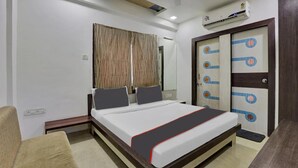 A hotel room with a bed, a bedside table, a chair, a wardrobe, and an air conditioning unit. - Hotel Alexa (Ahmedabad)