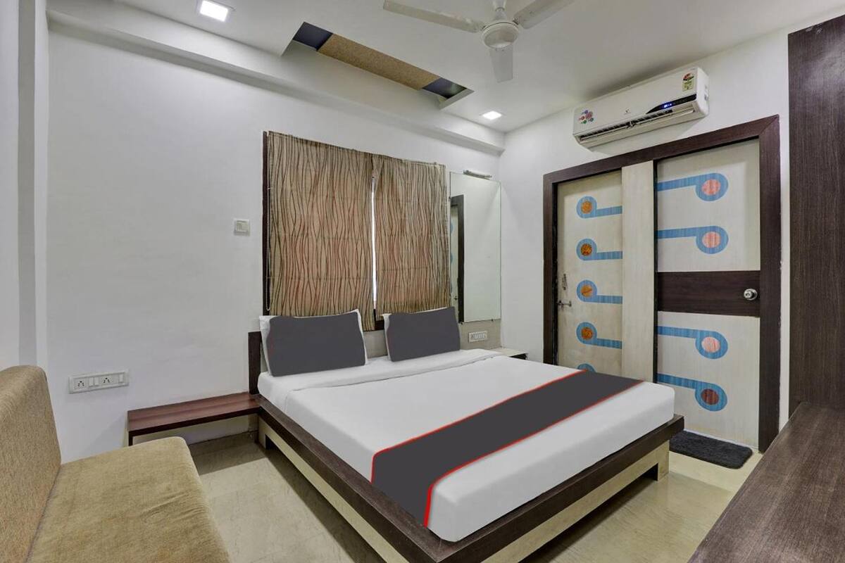 A hotel room with a bed, a bedside table, a chair, a wardrobe, and an air conditioning unit.