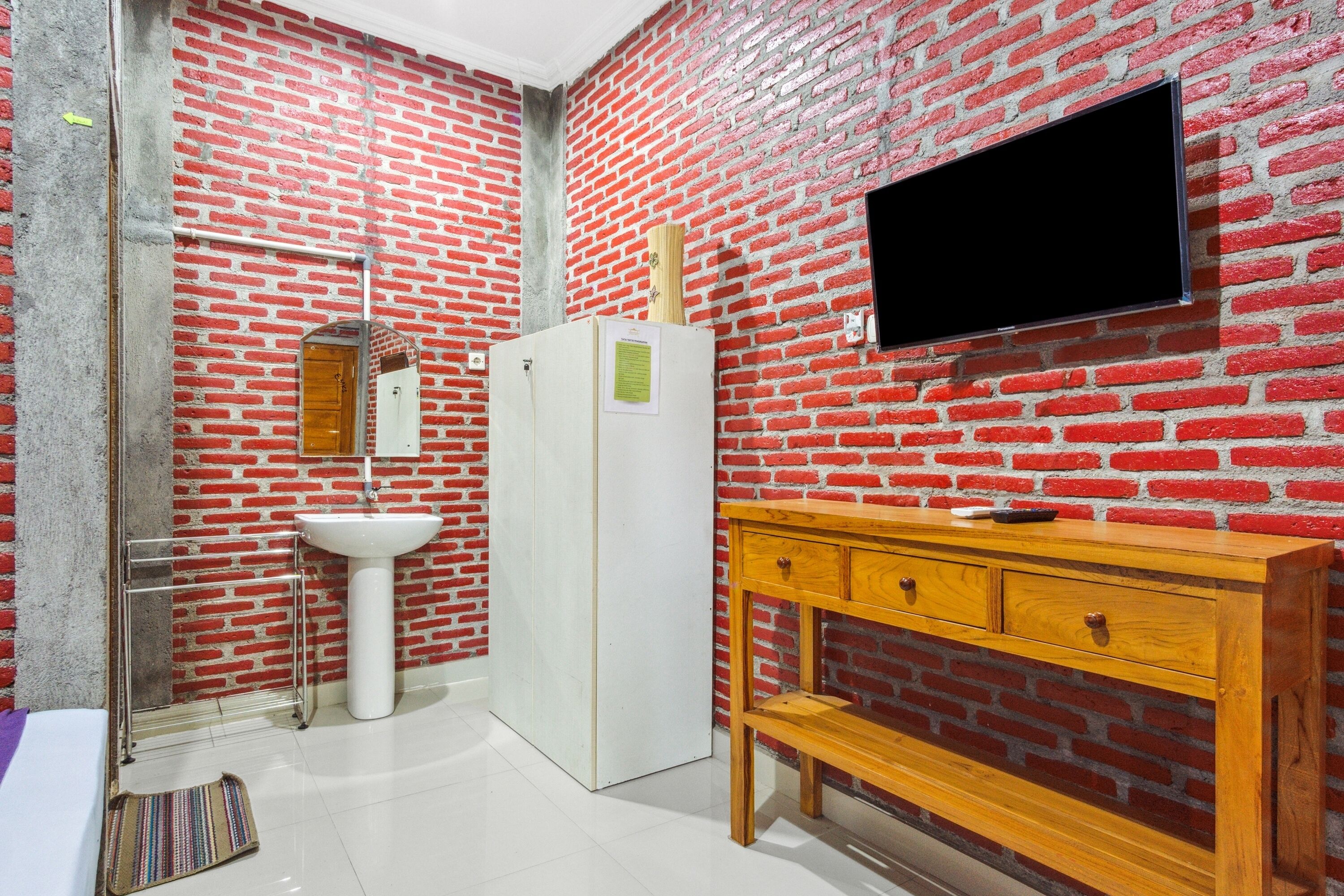 Une salle de bain avec un mur en briques rouges, un lavabo blanc, un miroir, un meuble-lavabo en bois et un téléviseur à écran plat fixé au mur.
