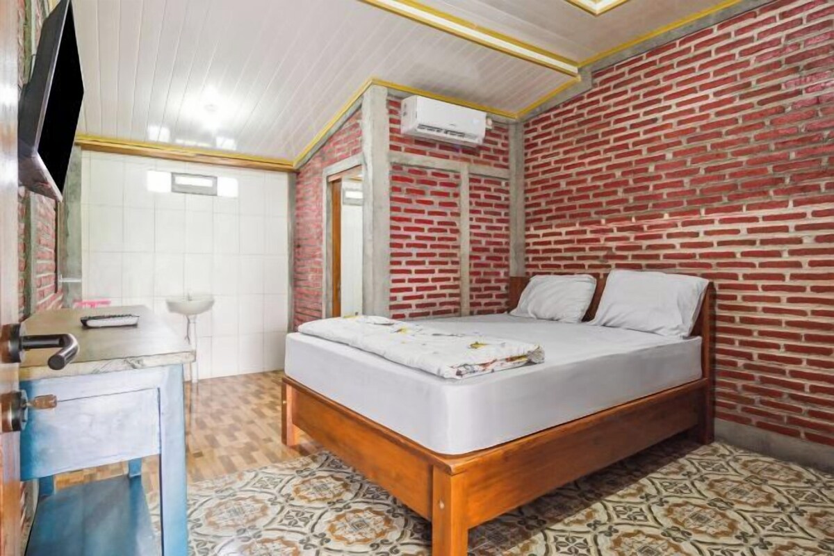 Um quarto com parede de tijolos, uma cama de madeira, colchão branco, mesinha de cabeceira pequena e piso estampado.