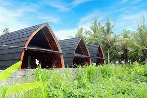 Een rij bungalows met puntdaken en open veranda's, omringd door weelderige begroeiing en palmbomen.