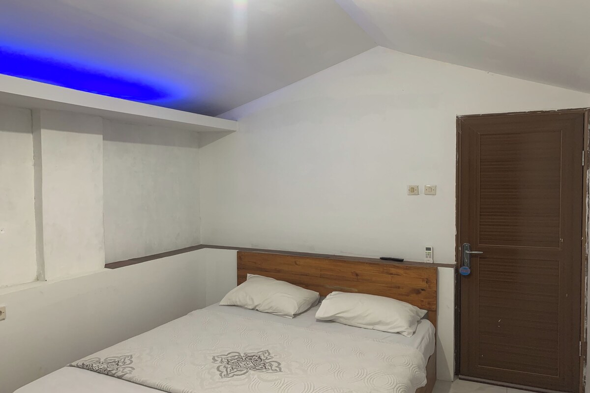 Quarto com cabeceira de madeira, cama com roupa de cama estampada, prateleira na parede e porta com puxador.