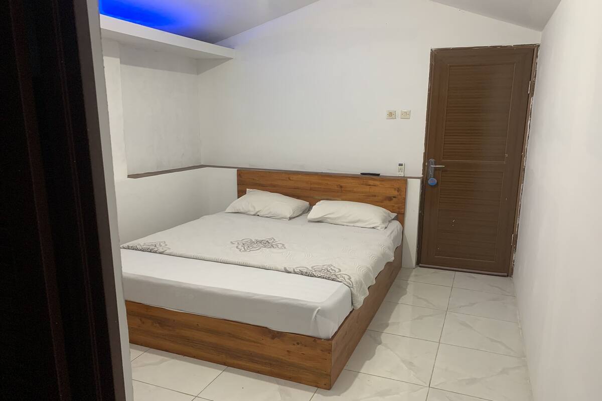 A bedroom with a wooden bed, white bedding, a closed door, and a white ceiling with a light fixture.