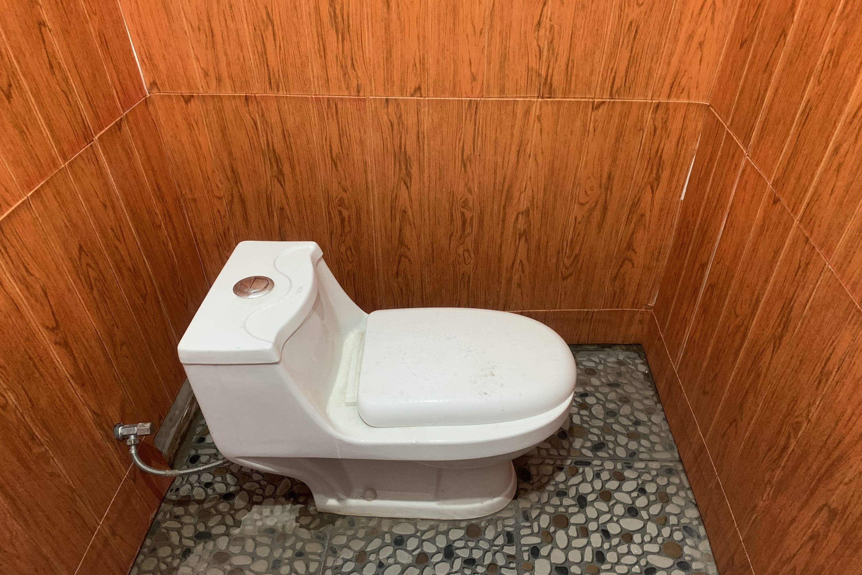 Un baño pequeño con paredes de madera, un inodoro blanco y suelo con estampado de piedras.