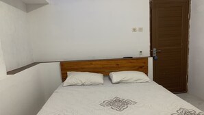 A single bed with a wooden headboard, two pillows, and a patterned bedspread in a room with white walls and a wooden door. - Mozza Guest House (Klaten)
