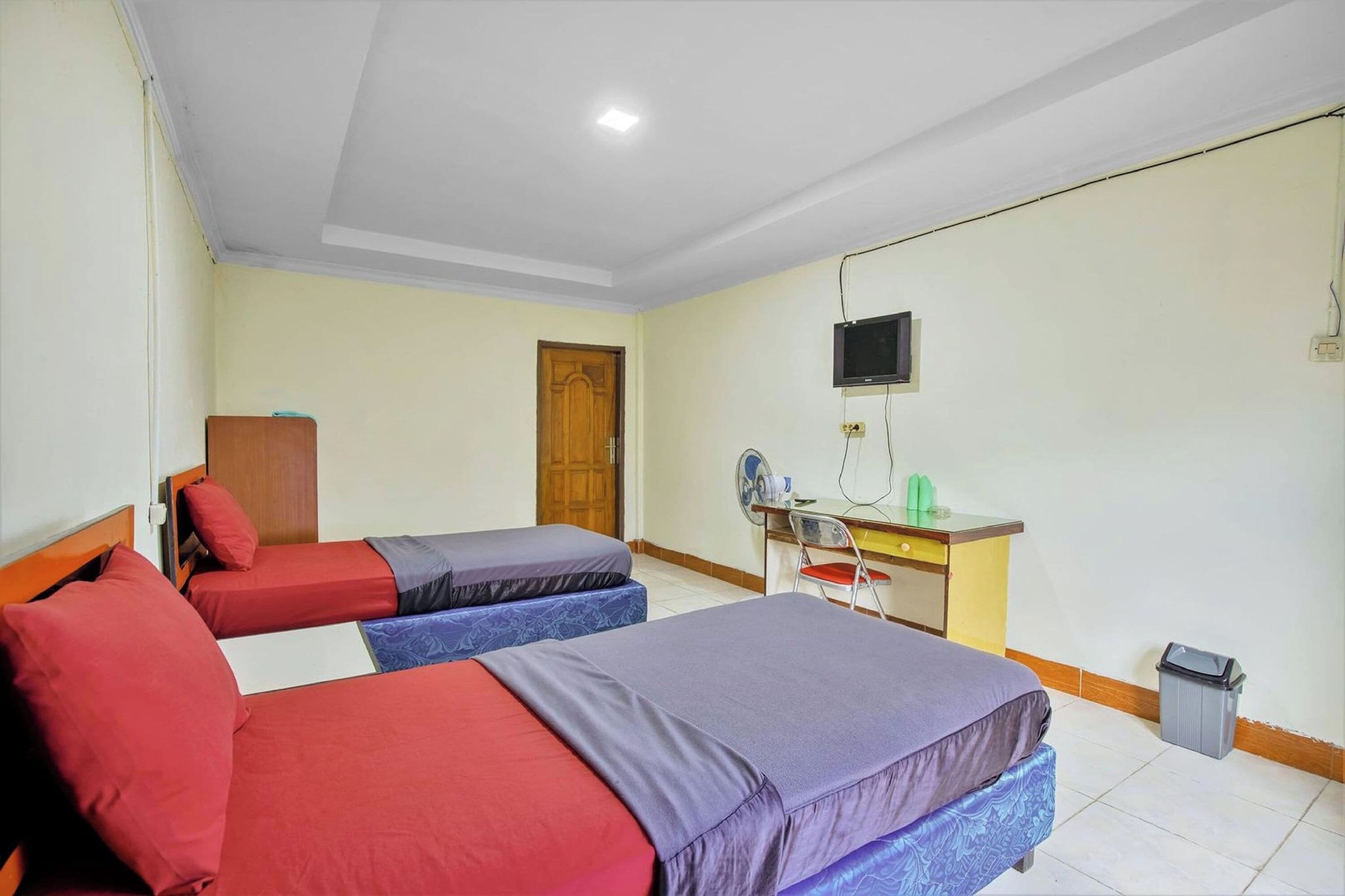 Habitación de hotel con dos camas, un escritorio con silla, un televisor y un cubo de basura.