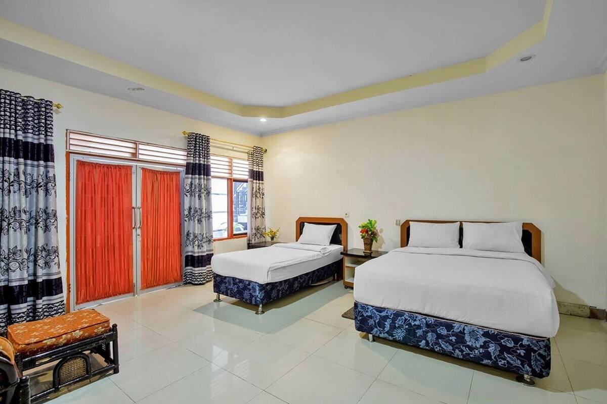 A hotel room with two beds, a wooden bench, a small table, and a window with curtains.