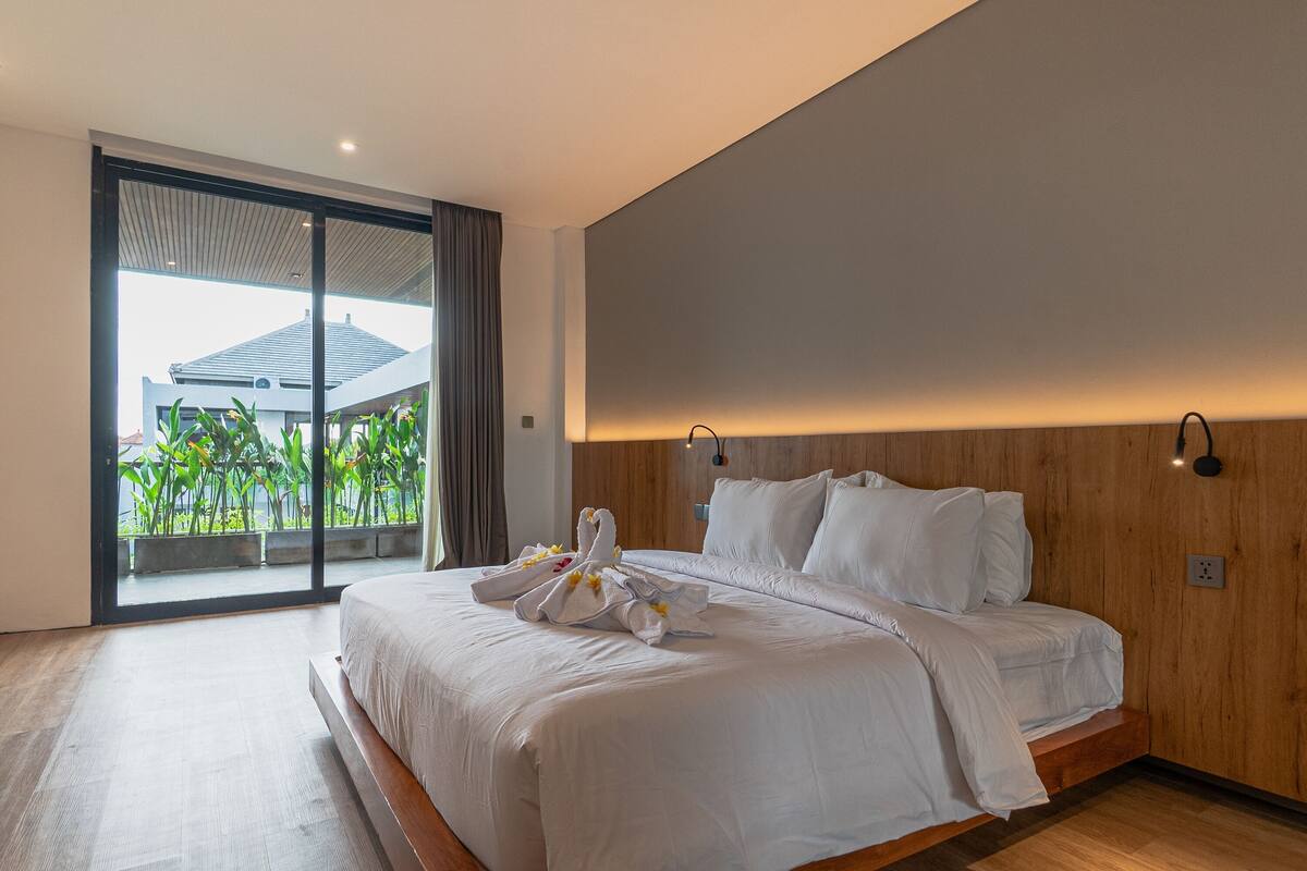 A modern hotel room with a large bed, a wooden headboard, and a balcony with plants.