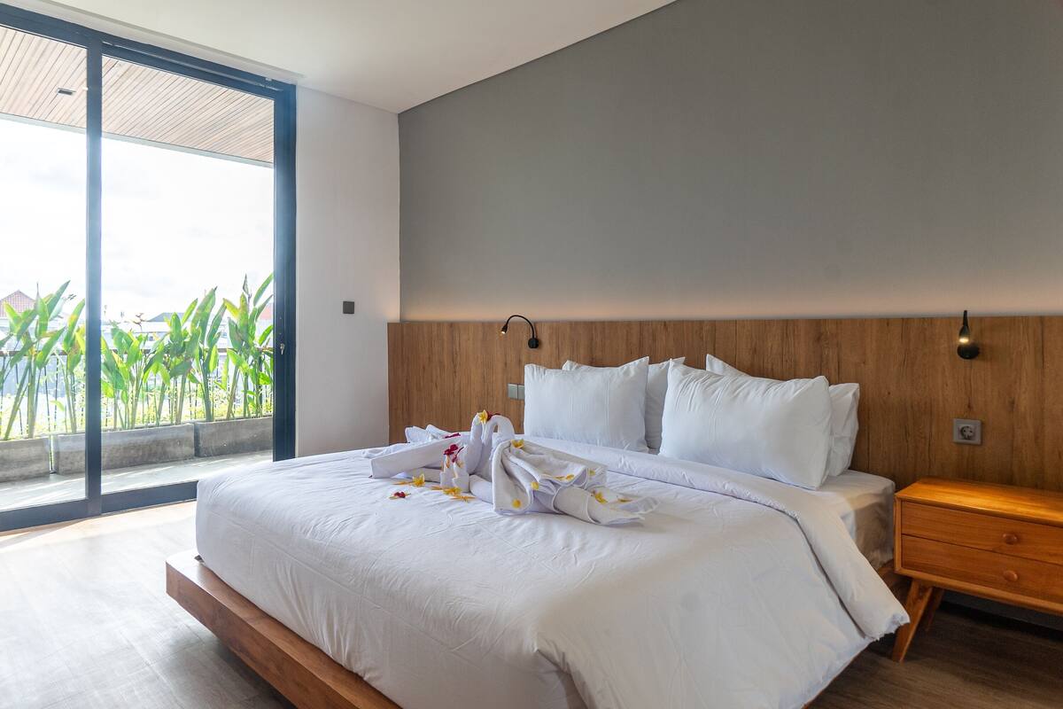 A modern hotel room with a large bed, wooden headboard, and a view of greenery through a sliding door.