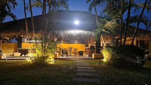 A well-lit outdoor area with a thatched-roof structure, surrounded by palm trees and a paved pathway leading to a seating area with tables and chairs. - Kanchira Resort (Pattaya)