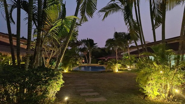 A well-lit pathway leading to a pool surrounded by palm trees and greenery, with a building in the background. - Kanchira Resort (Pattaya)