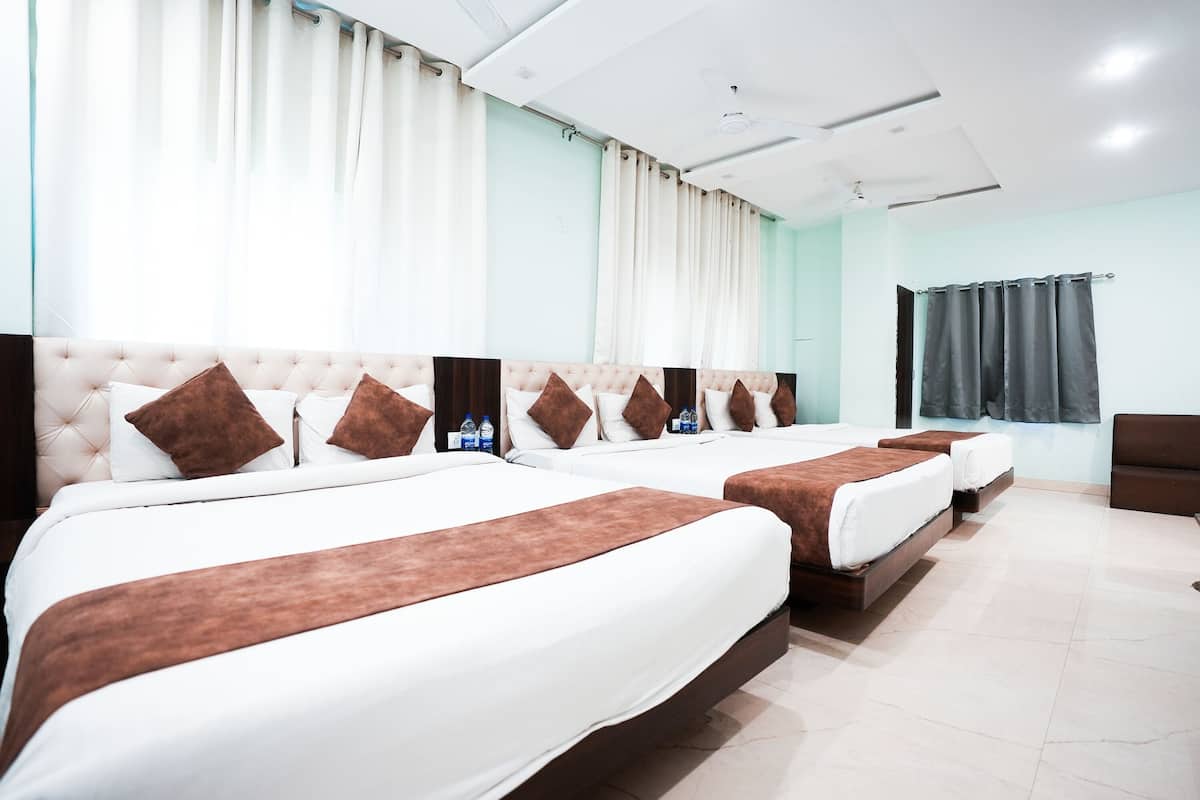 Quarto de hotel com três camas, cada uma com cabeceira marrom e roupa de cama branca. Cortinas nas janelas e uma TV de tela plana fixada na parede.