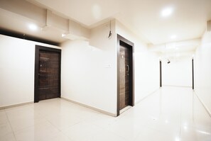 A hallway with two doors, one dark and one light, leading to rooms. The floor is tiled, and the walls are plain. - The White House (Pune)