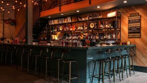 Una zona de bar con una barra larga, varios taburetes y una gran variedad de botellas y vasos.