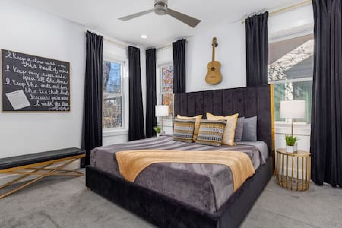 Une chambre moderne avec un grand lit, un ventilateur de plafond, une guitare accrochée au mur et une vue sur la ville par les fenêtres.