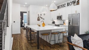 A modern kitchen with white cabinets, a central island with stools, pendant lighting, and a 'MUSIC ROW' sign. - Rooftop Deck, Patio & Garage - Walk to Restaurants (Nashville)