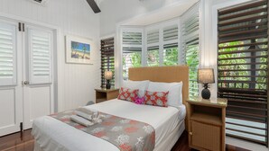 A neatly arranged bedroom with a bed, pillows, a bedside table with a lamp, and a window with shutters. - A Tropical Treat @ Sea Level / Port Douglas (PORT DOUGLAS)
