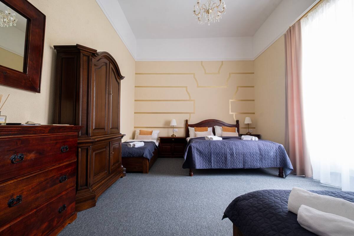 A hotel room with a large wooden dresser, two beds with blue bedding, a chandelier, and a patterned wall.