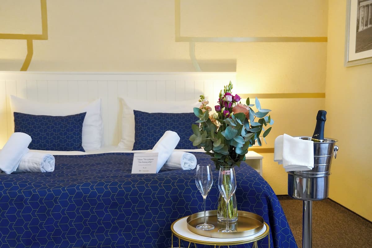A hotel room with a neatly made bed, a vase of flowers, two wine glasses, and a bucket with a towel.