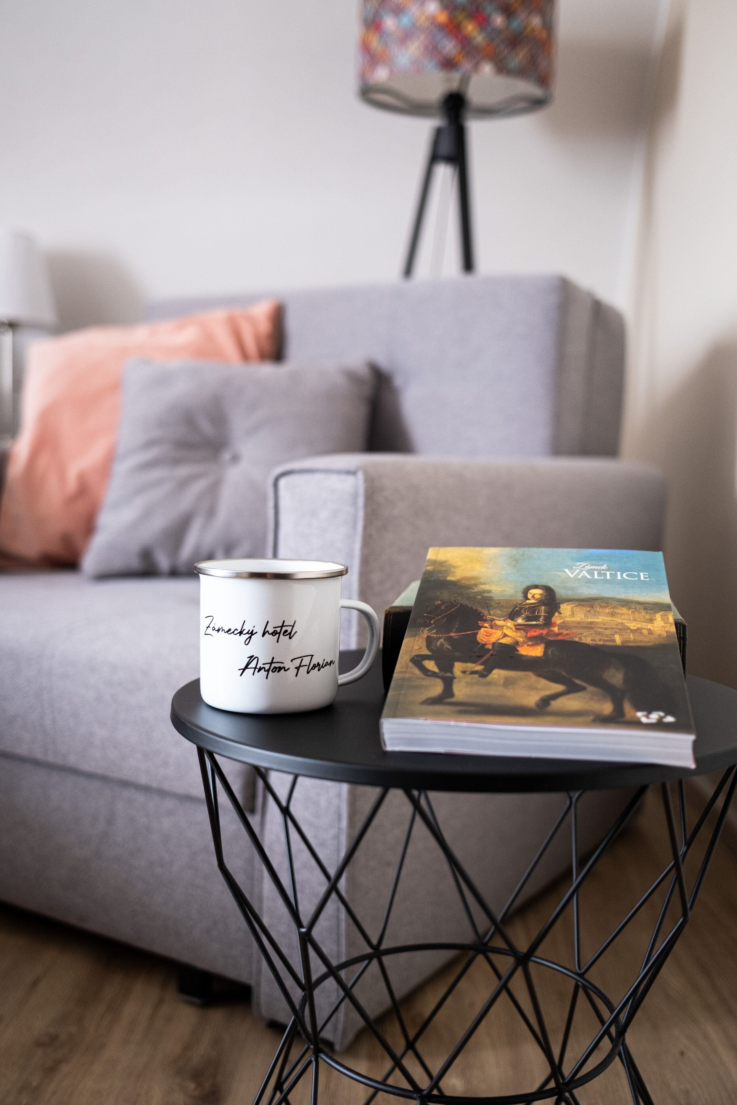A white mug with text on a black wire table, a book titled 'Valtice' beside it, and a grey sofa in the background.