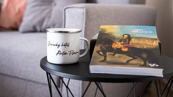 A white mug with text on a black wire table, a book titled 'Valtice' beside it, and a grey sofa in the background.