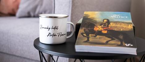 A white mug with text on a black wire table, a book titled 'Valtice' beside it, and a grey sofa in the background.